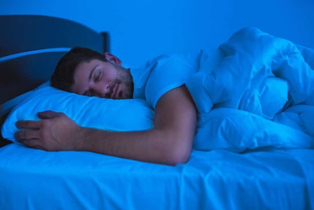 Maximizing Melatonin: Building a Nighttime Routine for Quality Sleep 13 Man peacefully sleeping in bed under a blue light, wrapped in a blanket with a relaxed expression. - was melatonin his trick?
