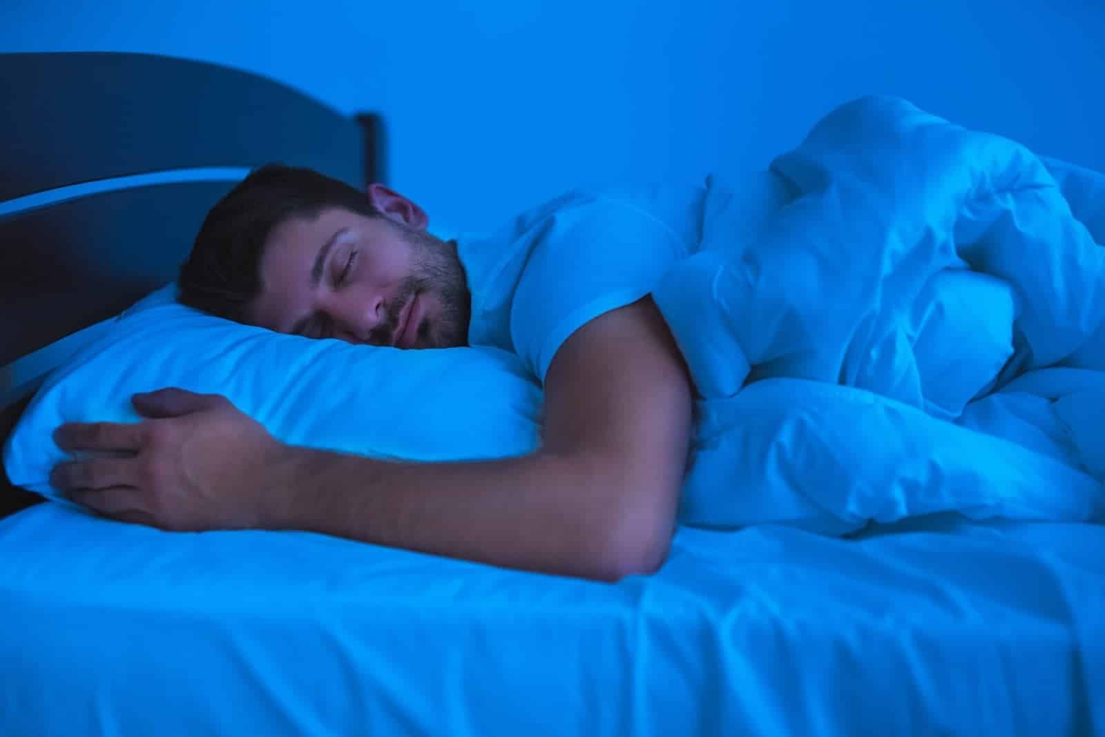 Man peacefully sleeping in bed under a blue light, wrapped in a blanket with a relaxed expression. - was melatonin his trick?