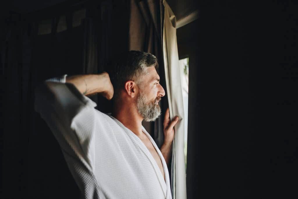 a man in a robe looking out a window thinking about melanin vs melatonin