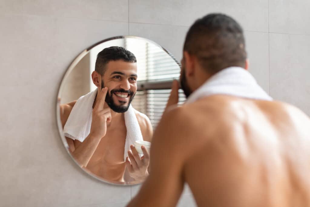 Is There a Vitamin F? The Truth About This Skincare Superstar 11 A smiling man looking wearing a towel in bathroom looking at the mirror putting a cream with proven Vitamin F on his cheek.