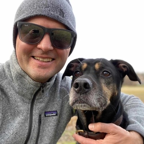 A man taking a selfie with his do in an open field. It's fall and cool outside.